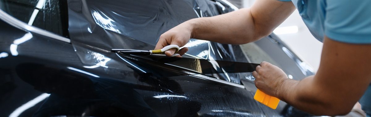 Male worker holds sheet of film, car tinting installation, tuning service. Mechanic applying vinyl tint on vehicle window in garage, tinted automobile glass